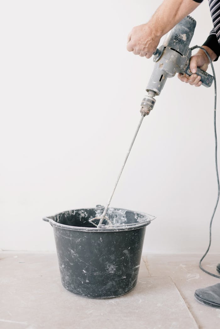 Worker using an electric drill to mix cement in a bucket during home renovation.